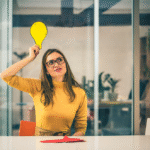 Mulher jovem com expressão pensativa segurando uma lâmpada de papel amarela acima da cabeça, simbolizando uma ideia ou insight, sentada em escritório moderno.